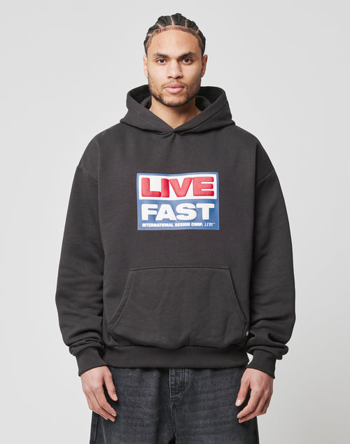 A man with braided hair wears an oversized black Live Fast Box Hooded hoodie by LIVE FAST DIE YOUNG, featuring a puffer print that spells LIVE FAST in red, white, and blue block letters. He stands against a plain light background.