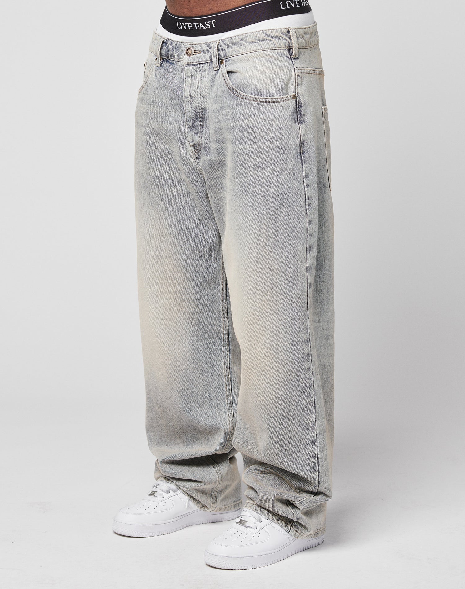 A person wears LFDY Baggy Denim in light-wash with wide, straight legs and a visible Flag Label, paired with white sneakers and black LIVE FAST waistband underwear, standing against a plain white background.