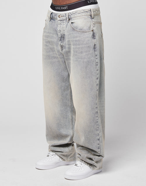 A person wears LFDY Baggy Denim in light-wash with wide, straight legs and a visible Flag Label, paired with white sneakers and black LIVE FAST waistband underwear, standing against a plain white background.