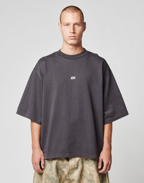 A person with short hair wears the LIVE FAST DIE YOUNG Live Fast Cropped Oversized Tee in dark gray with a white center logo, paired with light camo pants, standing against a plain light gray background.