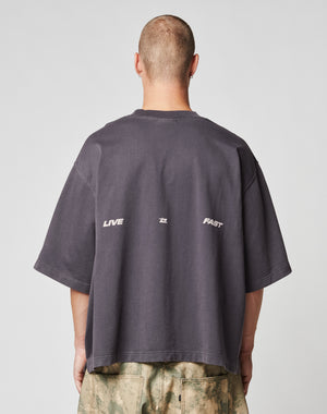 A person with a buzz cut, seen from behind, wears the LIVE FAST DIE YOUNG Live Fast Cropped Oversized Tee in dark gray with a bold backprint and star detail, paired with light green and beige tie-dye pants.