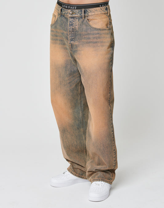 Someone wearing LFDY Baggy Denim by LIVE FAST DIE YOUNG—faded brown and blue, baggy fit with branded waistband—paired with white sneakers, standing against a plain white background.