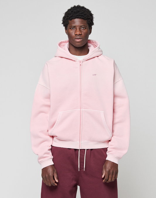 A person faces forward in a light pink LIVE FAST DIE YOUNG Basic Evidence Hooded Zip with an oversize fit and front pockets, paired with maroon pants, against a plain light gray background.