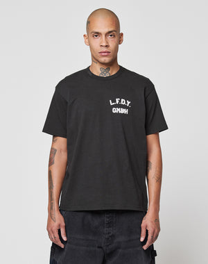 A person with a shaved head and visible tattoos wears the LFDY District Tee by LIVE FAST DIE YOUNG, featuring “L.F.D.Y. GMNH” front and back print, standing against a plain light gray background.