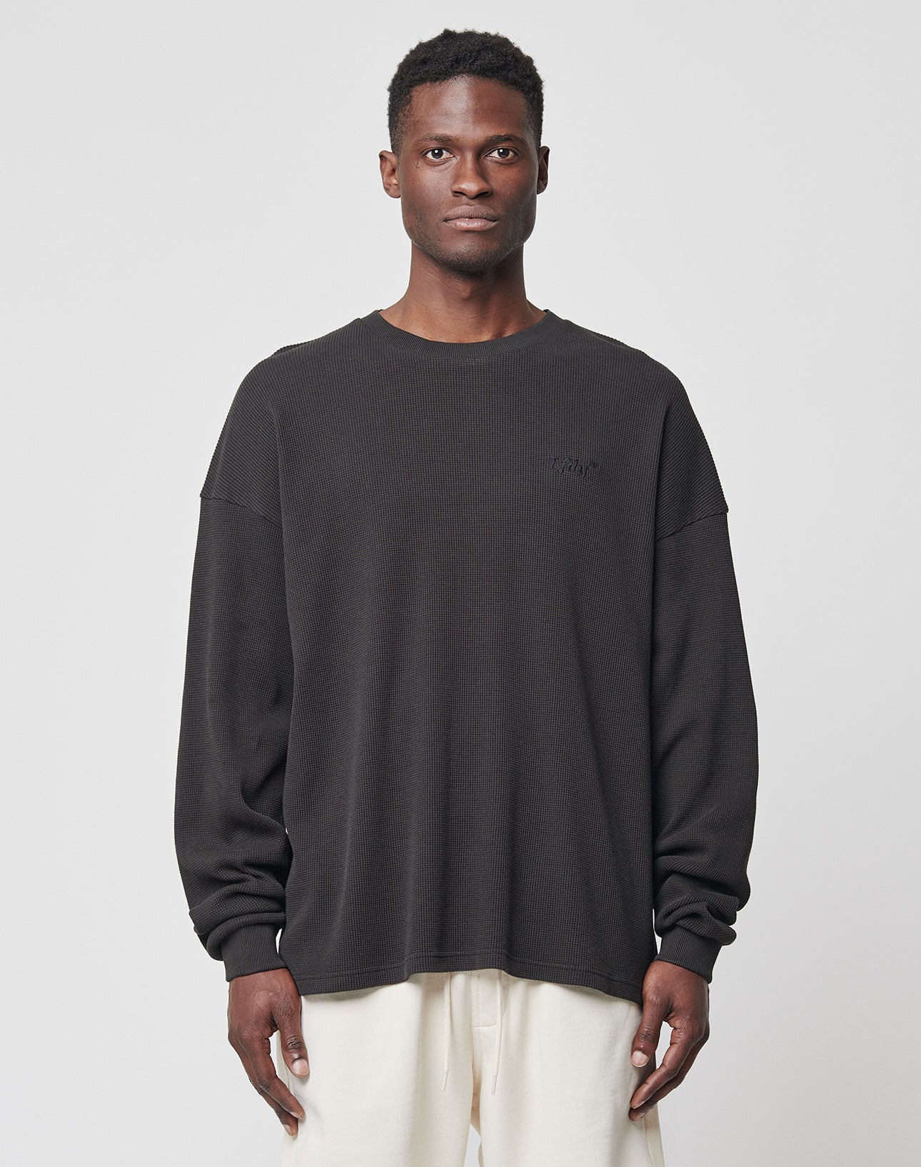 A man stands facing forward, wearing the LIVE FAST DIE YOUNG Basic Waffle Longsleeve in black with logo embroidery and light pants against a plain, light gray background.