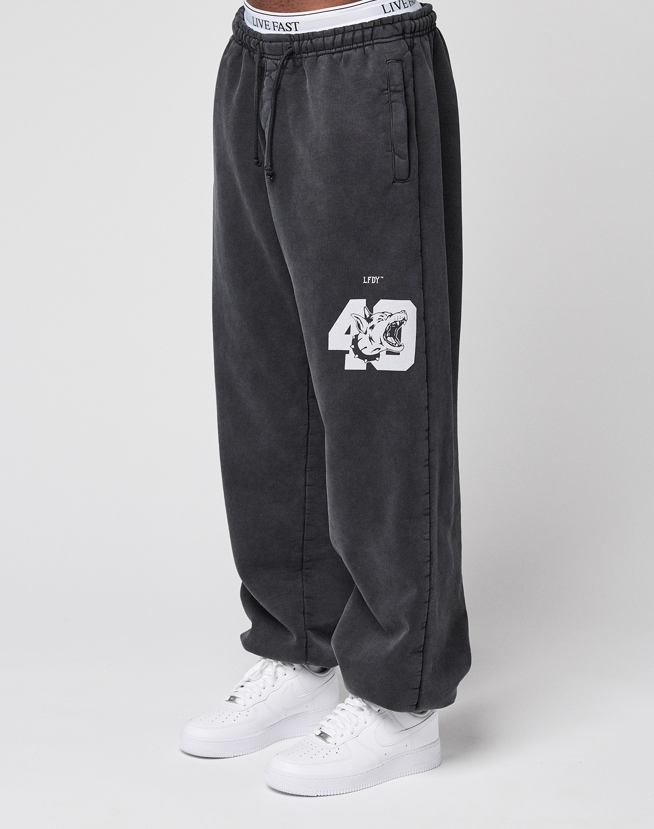A person in loose, dark gray LFDY 40 Wide Cuff Jogger sweatpants by LIVE FAST DIE YOUNG, featuring “49” and a tiger graphic on the left leg, side pockets, an oversize fit, white sneakers, and a visible "LIVE EAST" waistband.