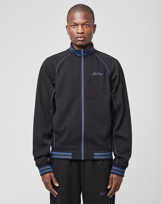 A person faces forward in a LIVE FAST DIE YOUNG Live Fast Tracksuit Jacket with Raglan sleeves, blue piping, and a small embroidered logo, paired with matching black track pants. The background is plain light gray.