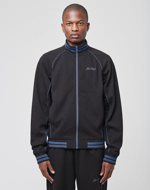 A person faces forward in a LIVE FAST DIE YOUNG Live Fast Tracksuit Jacket with Raglan sleeves, blue piping, and a small embroidered logo, paired with matching black track pants. The background is plain light gray.