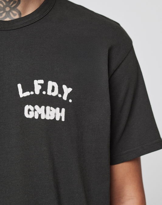 A person wears the LFDY District Tee by LIVE FAST DIE YOUNG, a regular fit black cotton t-shirt featuring white L.F.D.Y. GMBH text on the chest. Their tattooed neck is visible against a plain light background.