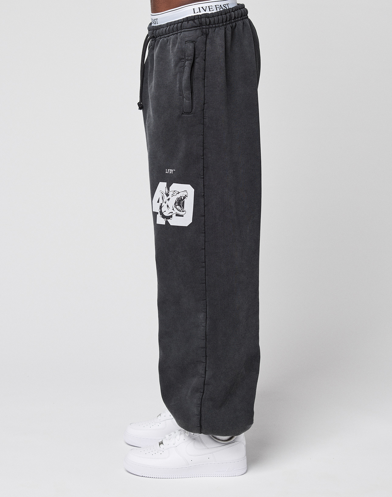 A person wears black LFDY 40 Wide Cuff Joggers by LIVE FAST DIE YOUNG with an oversize fit, side pockets, white '40' and tiger graphic, and a white LIVE FAST waistband, paired with white sneakers, standing sideways against a plain background.