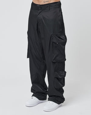 A person wears LIVE FAST DIE YOUNG Nylon Cargo pants—loose black cargo style with multiple pockets—and white sneakers, standing against a plain white background. Only the lower torso and legs are shown.