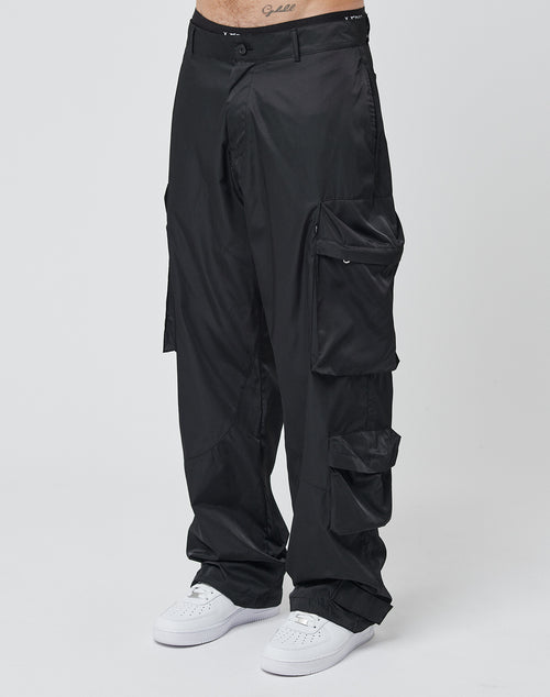 A person wears LIVE FAST DIE YOUNG Nylon Cargo pants—loose black cargo style with multiple pockets—and white sneakers, standing against a plain white background. Only the lower torso and legs are shown.