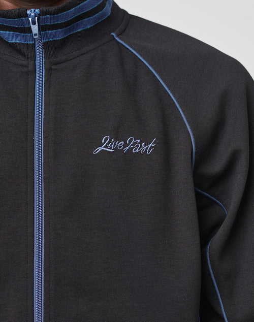 A close-up of a person wearing the Live Fast Tracksuit Jacket by LIVE FAST DIE YOUNG, featuring blue piping, a Ripp-Kragen, a blue and white striped collar, and Live Fast embroidered in white on the chest.