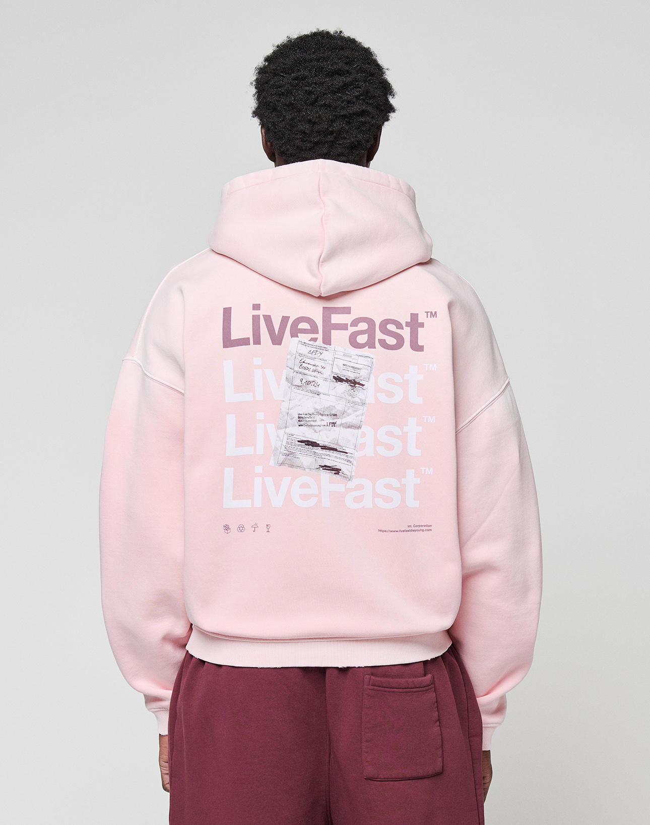 A person wears a LIVE FAST DIE YOUNG Basic Evidence Hooded Zip in oversize fit, light pink with white and purple "LiveFast" print and crumpled paper graphic on the back, paired with maroon pants.