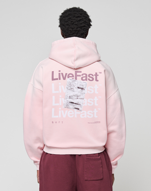 A person wears a LIVE FAST DIE YOUNG Basic Evidence Hooded Zip in oversize fit, light pink with white and purple "LiveFast" print and crumpled paper graphic on the back, paired with maroon pants.