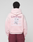 A person wears a LIVE FAST DIE YOUNG Basic Evidence Hooded Zip in oversize fit, light pink with white and purple "LiveFast" print and crumpled paper graphic on the back, paired with maroon pants.