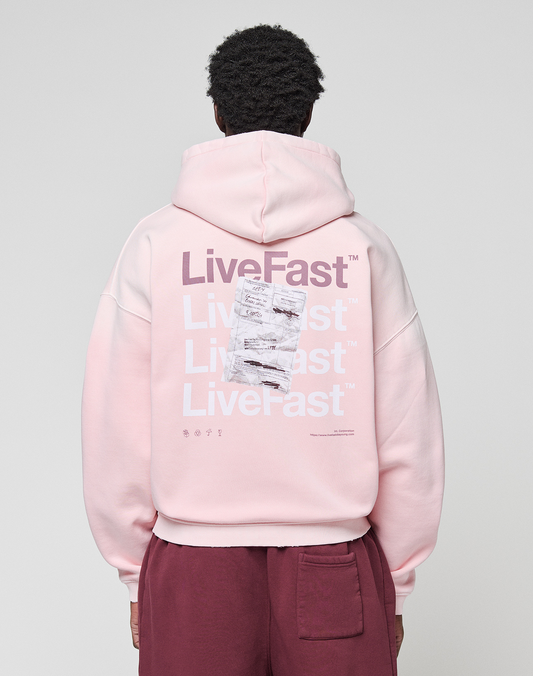 A person wears a LIVE FAST DIE YOUNG Basic Evidence Hooded Zip in oversize fit, light pink with white and purple "LiveFast" print and crumpled paper graphic on the back, paired with maroon pants.