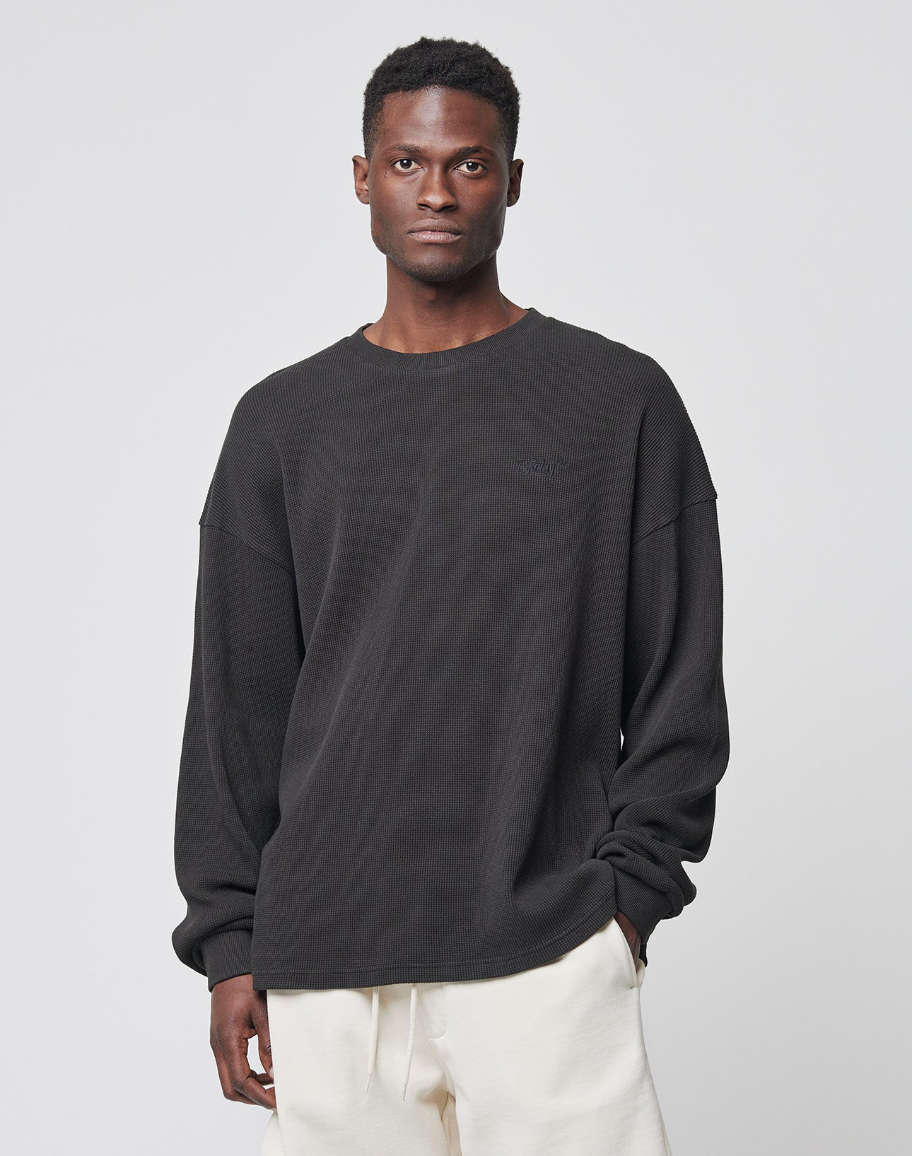 A man stands against a light gray background, wearing the LIVE FAST DIE YOUNG Basic Waffle Longsleeve in black (100% Baumwolle) and off-white pants, with one hand in his pocket and a neutral expression.