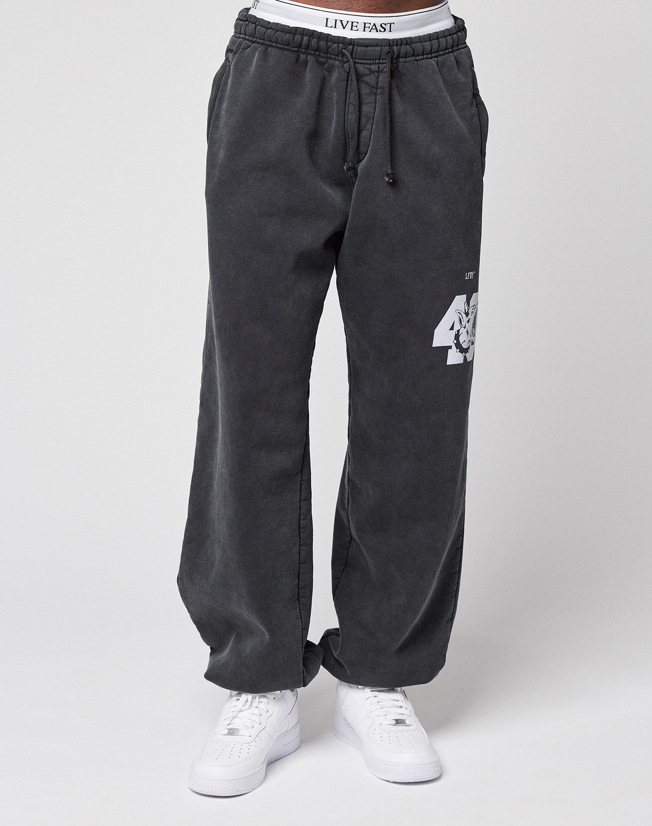 A person wears LFDY 40 Wide Cuff Joggers by LIVE FAST DIE YOUNG, featuring an oversize dark gray cotton fit, white '47' and tiger graphic on the left leg, side pockets, a white "LIVE FAST" waistband, and white sneakers. Plain light gray background.