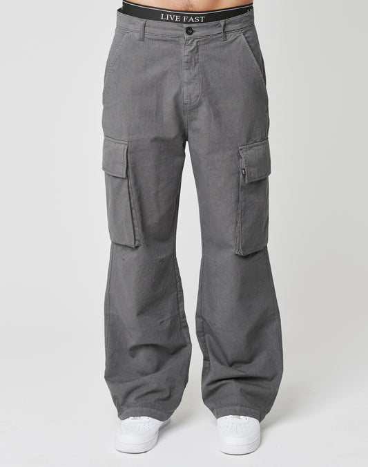 A person wears the LIVE FAST DIE YOUNG Wide Cargo—a gray, wide fit pant with Cargo-Taschen and side pockets—styled with white sneakers. The black waistband reads LIVE FAST. Theyre standing against a plain white background.