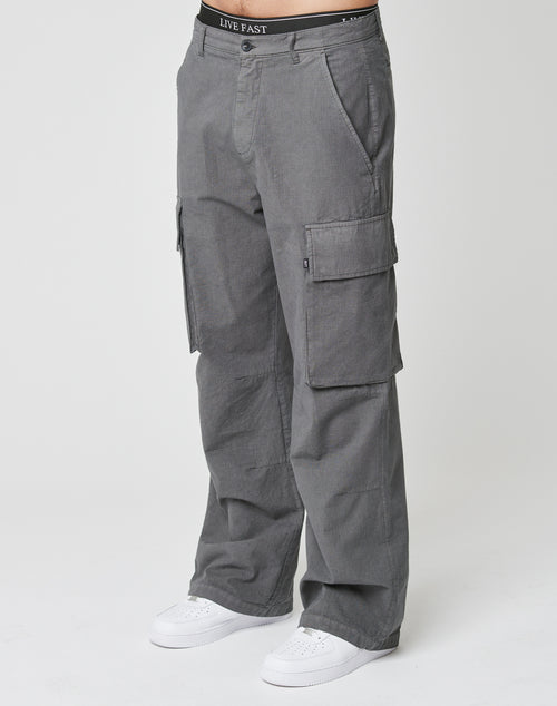 A person in LIVE FAST DIE YOUNG Wide Cargo gray pants with large cargo pockets, white sneakers, and a visible LIVE FAST waistband stands against a plain white background.