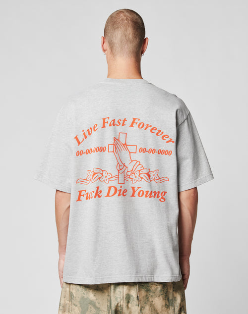 A person with a shaved head wears the LIVE FAST DIE YOUNG LFDY Praying Tee, an oversize gray t-shirt with a bold back print above praying hands, a cross, and flowers. Camouflage pants complete the look.
