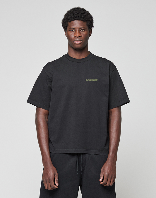 A person faces forward, wearing the LIVE FAST DIE YOUNG Basic 210 Tee—an oversized black short-sleeve shirt made from 100% cotton with "LiveFast" in small green letters on the left chest—and black pants, against a plain light gray background.