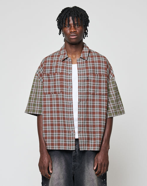 A young man with short dreadlocks wears the LIVE FAST DIE YOUNG LFDY Light Flannel—an oversize fit, short-sleeve, plaid button-up in brown, green, and grey—over a white tee and dark jeans against a plain light background.