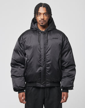 A man with braided hair wears the LIVE FAST DIE YOUNG LFDY Winter Bomber Jacket in black with an oversized fit and hood, paired with black pants, standing facing forward against a plain light gray background.