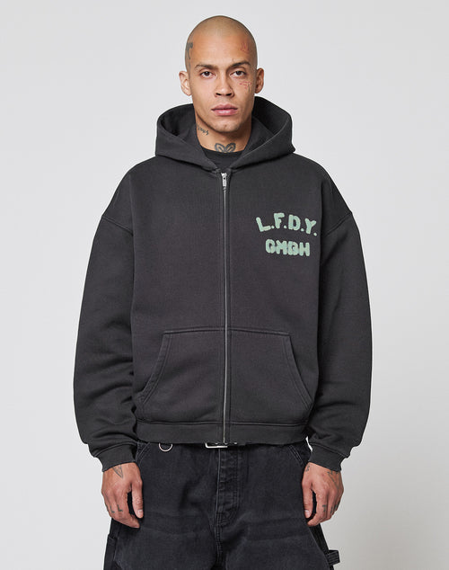 A man with a shaved head wears the Live Fast District Hooded Zip by LIVE FAST DIE YOUNG, an oversized black zip-up jacket with light blue LFDY branding on the chest, standing against a plain white background.