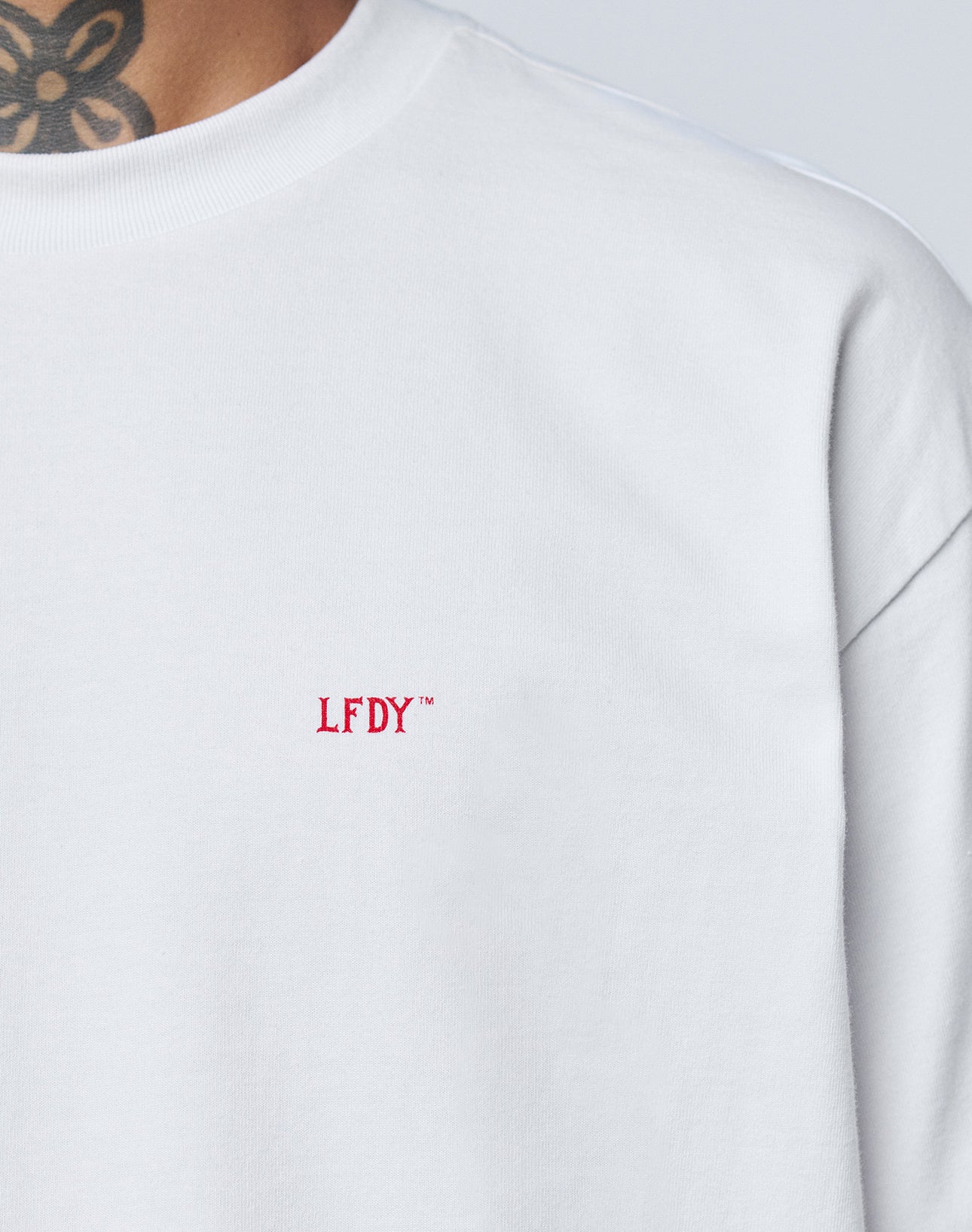 Close-up of a person wearing the Basic Evidence Tee by LIVE FAST DIE YOUNG, an oversize fit white shirt with small red LFDY™ embroidery on the chest. Made from 100% Baumwolle, paired with a black neck tattoo.