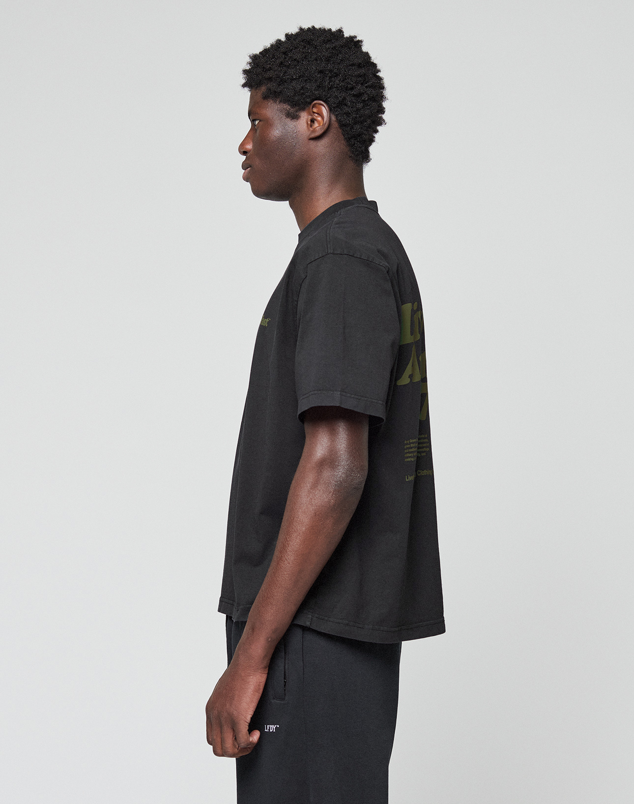 A person with short curly hair stands in profile against a plain background, wearing the LIVE FAST DIE YOUNG Basic 210 Tee in black. The oversize fit t-shirt features front and back graphics and text on the sleeve, paired with black pants.