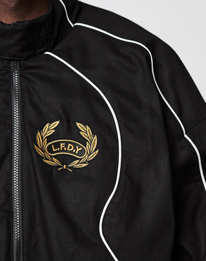 Close-up of the black LFDY Canvas Jacket by LIVE FAST DIE YOUNG, featuring white piping, multi-logo embroidery including a gold laurel wreath, and L.F.D.Y on the left chest.