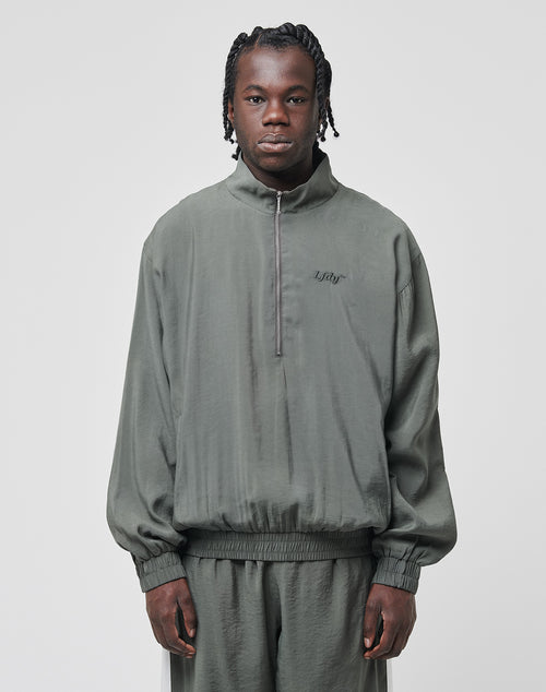 A man with braided hair faces forward, wearing a muted green LIVE FAST DIE YOUNG LFDY Light Halfzip jacket and matching pants with a metal zipper. The background is plain light gray.