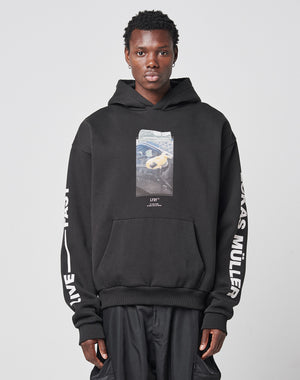 A person wearing the Mirror Hooded oversize black hoodie by LIVE FAST DIE YOUNG, featuring a rectangular front print and white LIVE FAST and LUKAS MÜLLER text on the sleeves, stands against a plain light background.