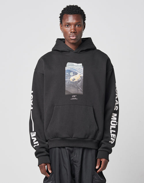 A person wearing the Mirror Hooded oversize black hoodie by LIVE FAST DIE YOUNG, featuring a rectangular front print and white LIVE FAST and LUKAS MÜLLER text on the sleeves, stands against a plain light background.