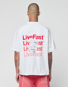 Seen from behind, a person with a shaved head wears the LIVE FAST DIE YOUNG Basic Evidence Tee—a white, 100% Baumwolle oversize T-shirt with red LiveFast prints on front and back—paired with pink shorts against a plain light background.