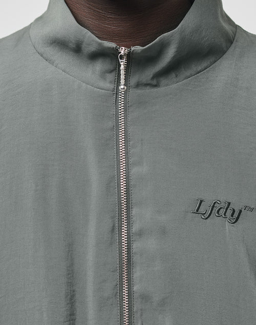 Close-up of the LIVE FAST DIE YOUNG LFDY Light Halfzip in light gray, featuring a metallic zip and stand-up collar. “LFDY” is embroidered on the left chest in dark thread; only the wearer’s neck and upper chest are visible.