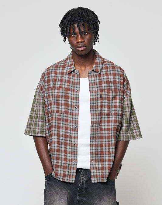 A young man with short dreadlocks wears the LIVE FAST DIE YOUNG LFDY Light Flannel—an oversized brown and green plaid shirt—over a white tank top and dark jeans, standing against a plain light-grey background.