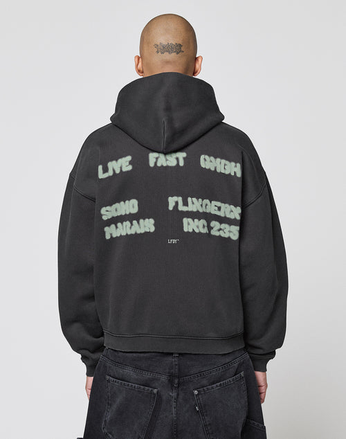 A person with a shaved head and tattoo wears the LIVE FAST DIE YOUNG Live Fast District Hooded Zip in black, featuring bold white spray-paint style text on the back for a signature streetwear look.