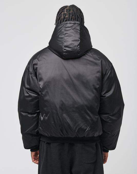 A person with braided hair, seen from the back against a plain light gray background, is wearing an oversize fit shiny black LFDY Winter Bomber Jacket by LIVE FAST DIE YOUNG with a hood.