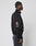 A person in profile wears the LFDY Canvas Jacket by LIVE FAST DIE YOUNG—black with white piping, multi-logo patches and flag details, plus elastic cuffs. They have braided hair and pair it with faded black jeans.