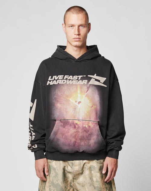 A person with short hair wears the LFDY Angel Hooded by LIVE FAST DIE YOUNG—an oversized black hoodie with a bold graphic—styled with tan camouflage pants against a plain light gray background.