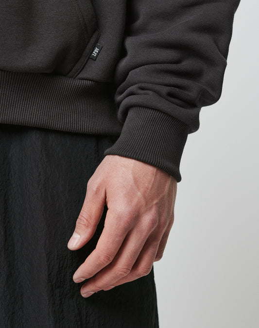 A close-up of a person’s left hand and wrist, wearing black textured pants and the oversized Hit and Run Hooded sweatshirt by LIVE FAST DIE YOUNG, featuring ribbed fabric and a small fabric tag near the waistband.