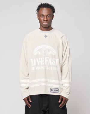 A person with short, twisted hair wears the LFDY Hockey Jersey by LIVE FAST DIE YOUNG, an oversize beige hockey-style jersey featuring front and sleeve prints, standing against a plain light background.
