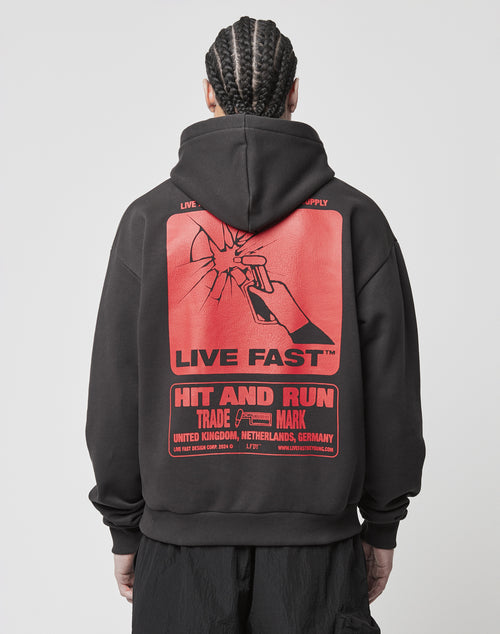 A person with braided hair wears the LIVE FAST DIE YOUNG Hit and Run Hooded—a black oversized hoodie with a large cracked backprint of a hand lighting a cigarette, bold “LIVE FAST” and “HIT AND RUN” text, plus smaller details.