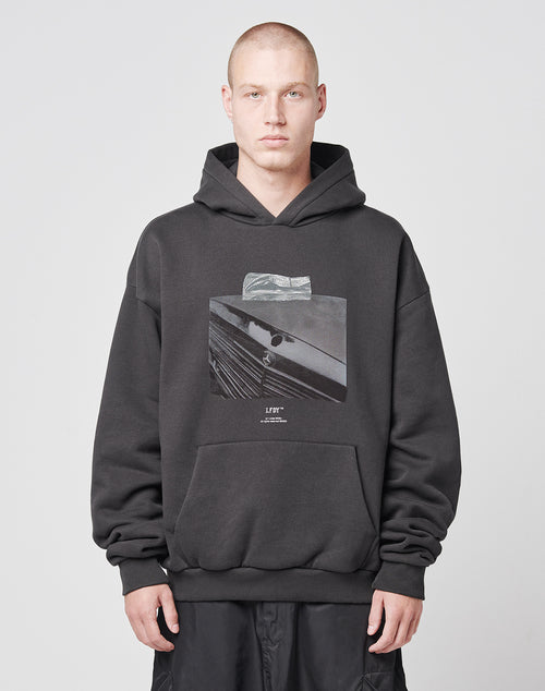 A person with a shaved head wears the LIVE FAST DIE YOUNG Benz Hooded, an oversize black hoodie with bold frontprint, paired with black pants against a plain light background.