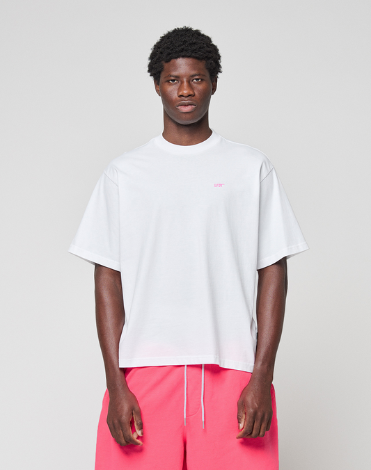 A person faces forward, wearing a loose white LIVE FAST DIE YOUNG Basic Evidence Tee in Oversize Fit and bright pink shorts against a plain light gray background.