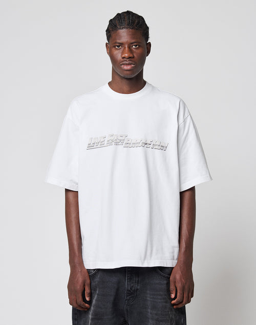A young man with braided hair faces forward, wearing the LIVE FAST DIE YOUNG LFDY Europe Run Tee—a loose, oversized white t-shirt with embossed text—paired with dark jeans against a plain light gray background.