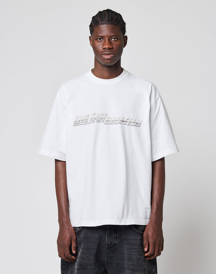 A young man with braided hair faces forward, wearing the LIVE FAST DIE YOUNG LFDY Europe Run Tee—a loose, oversized white t-shirt with embossed text—paired with dark jeans against a plain light gray background. | model
